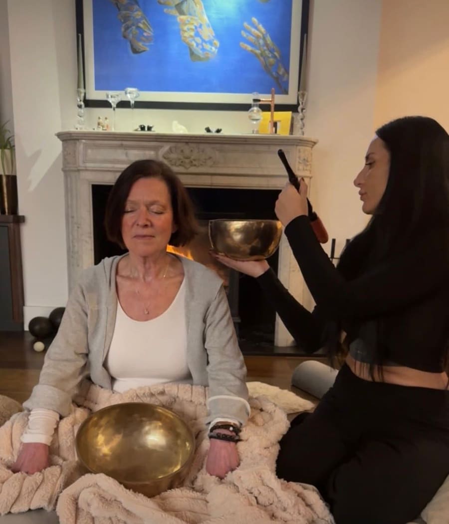Gem playing a Tibetan singing bowl beside a client resting under a blanket during a private session